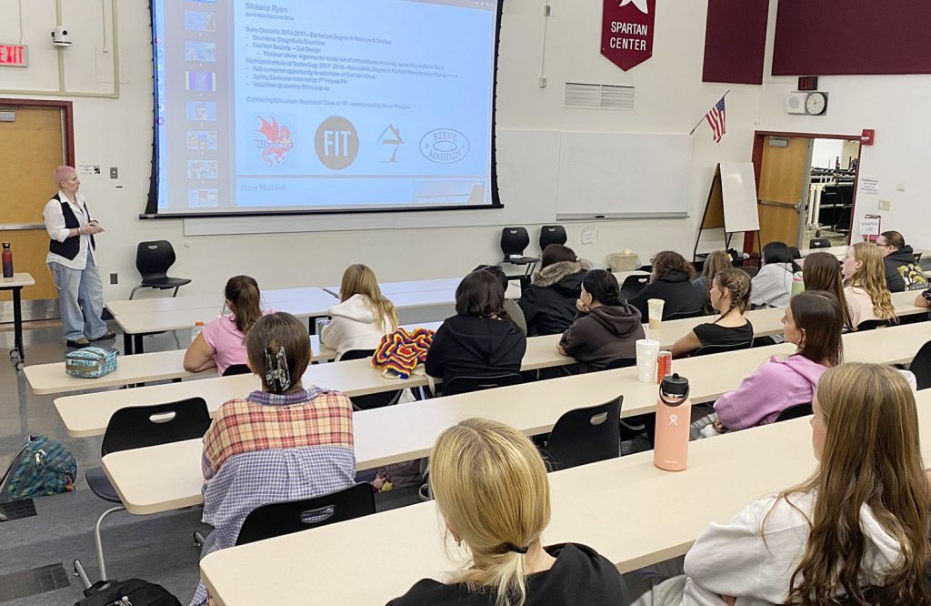 High school students sit at long tables inside a classroom and listen to a guest speaker who is standing at the front of the room